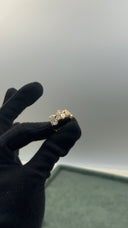 A gold ring with clear gemstones held between a hand wearing a black glove, set against a neutral gray background.