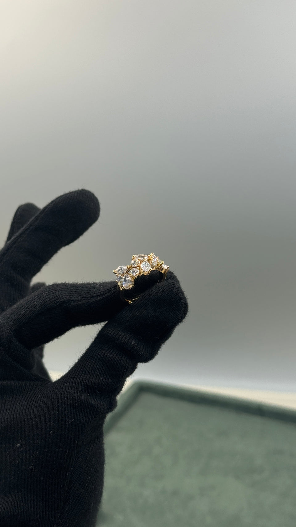 A gold ring with clear gemstones held between a hand wearing a black glove, set against a neutral gray background.
