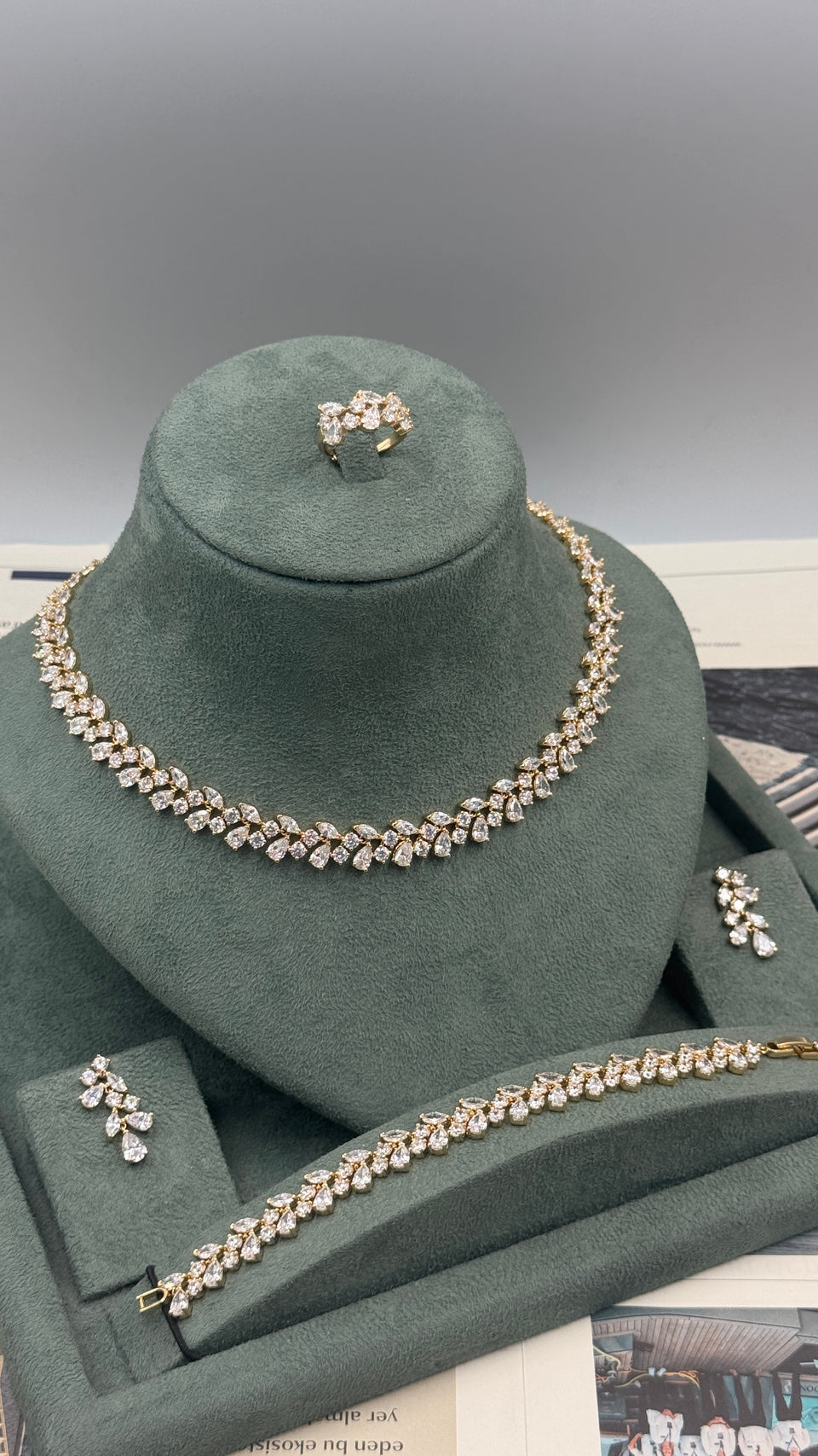 A gold-tone jewelry set with clear rhinestones displayed on a dark green velvet jewelry stand. The set includes a choker necklace, a matching ring, a bracelet, and two matching earrings.