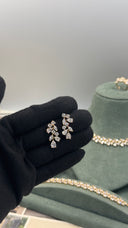 A close-up of a hand wearing a black glove holding a pair of gold-toned leaf-shaped stud earrings with clear stones. In the background, a green jewelry display bust holds matching gold-toned necklaces and a ring.