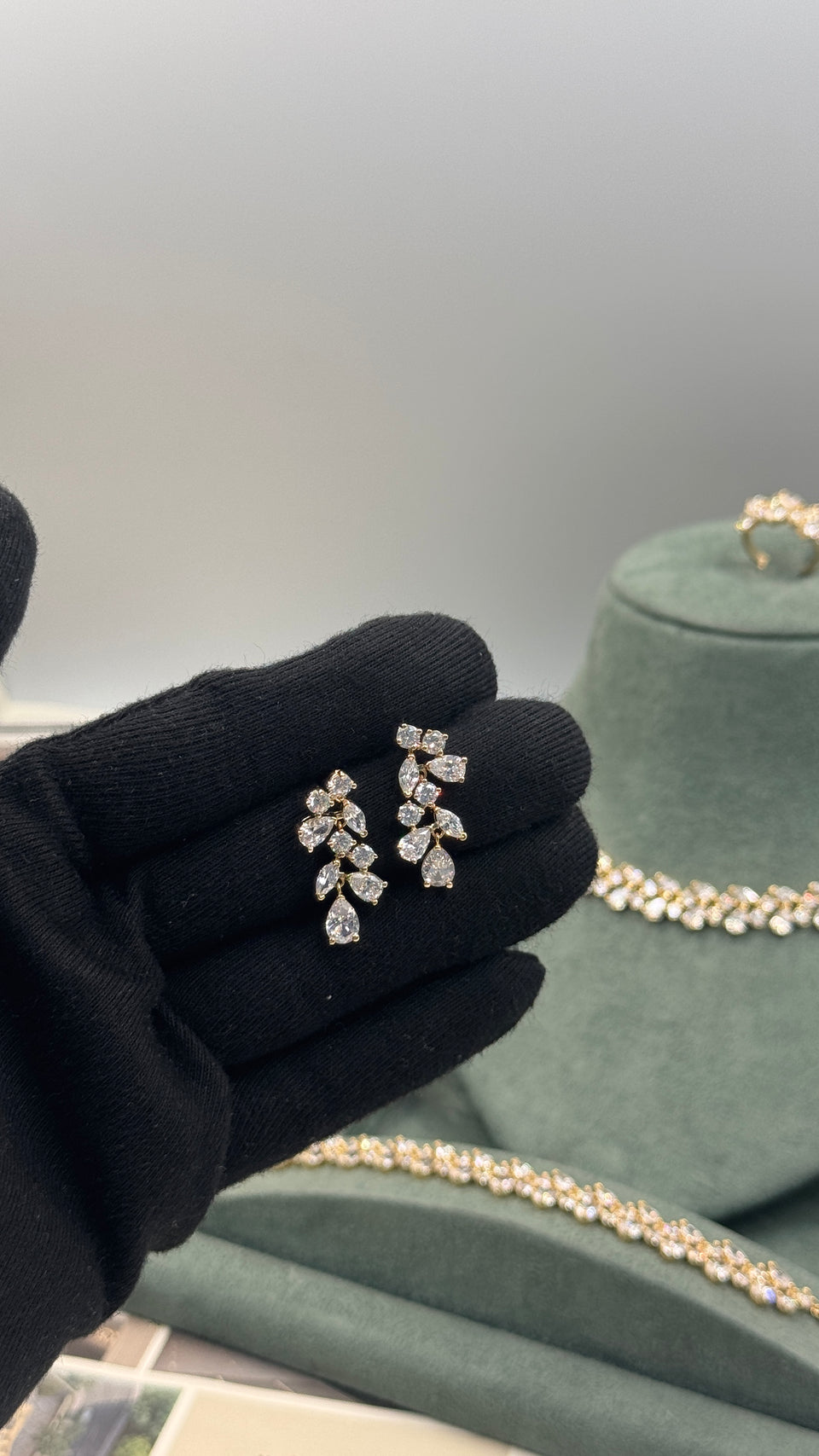A close-up of a hand wearing a black glove holding a pair of gold-toned leaf-shaped stud earrings with clear stones. In the background, a green jewelry display bust holds matching gold-toned necklaces and a ring.