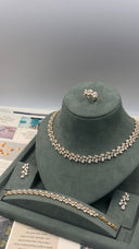 A jewelry set displayed on a dark green velvet mannequin bust, including a necklace, bracelet, ring, and earrings, all featuring clear rhinestones with gold-toned settings. The set is arranged on a gray tray with printed papers underneath.
