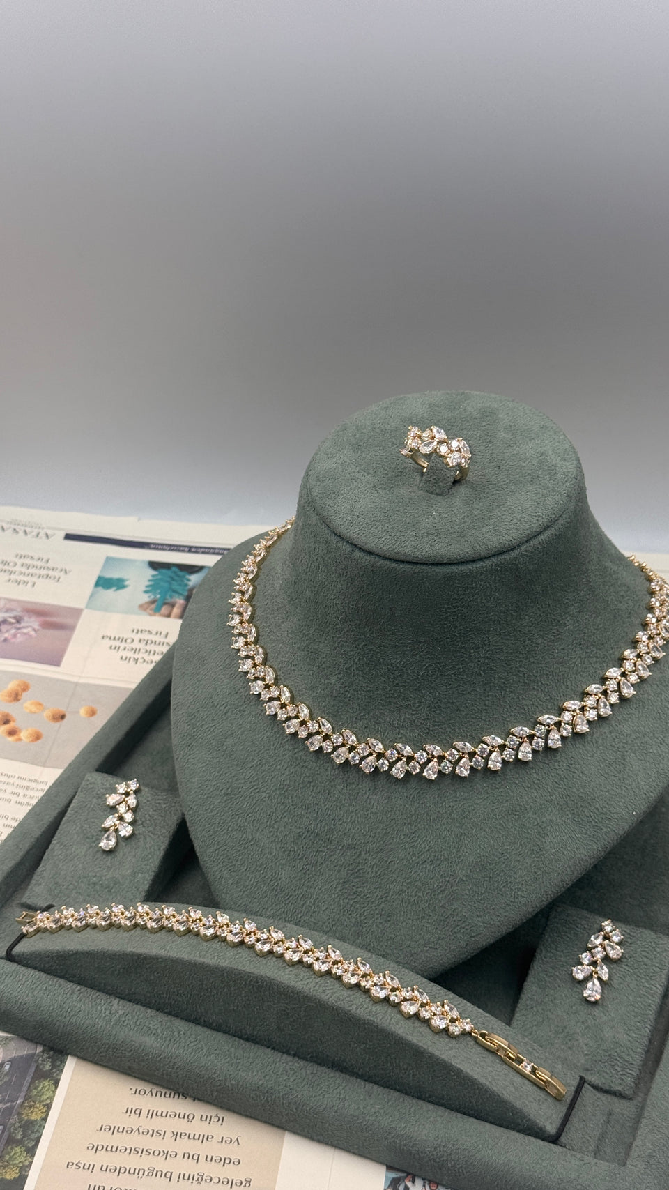 A jewelry set displayed on a dark green velvet mannequin bust, including a necklace, bracelet, ring, and earrings, all featuring clear rhinestones with gold-toned settings. The set is arranged on a gray tray with printed papers underneath.