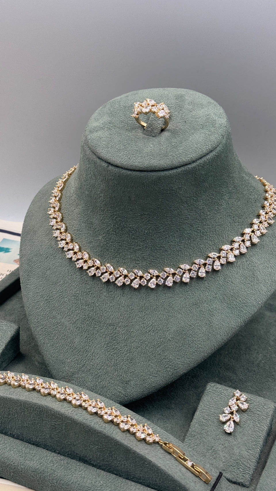 A jewelry set displayed on a dark green velvet mannequin bust, including a necklace, bracelet, ring, and matching pin. The jewelry features clear gemstones set in a yellow gold-tone metal.