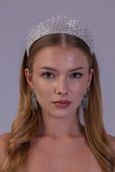 A woman with long blonde hair wears a sparkling silver crystal tiara and matching dangling crystal earrings against a solid gray studio background.