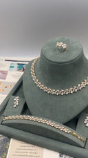 A jewelry set displayed on a dark green velvet stand, including a necklace, bracelet, ring, and earrings with clear rhinestones in a silver-tone setting, photographed against a neutral gray background.