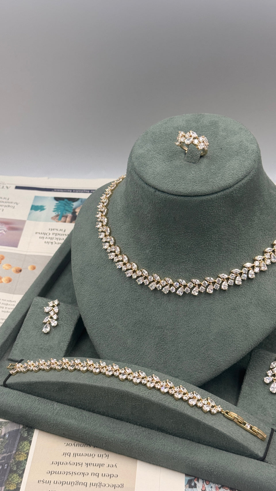 A jewelry set displayed on a dark green velvet stand, including a necklace, bracelet, ring, and earrings with clear rhinestones in a silver-tone setting, photographed against a neutral gray background.