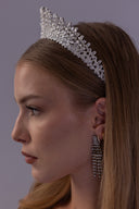 A woman in profile wearing a sparkling silver tiara headband and matching dangling earrings against a plain gray background.