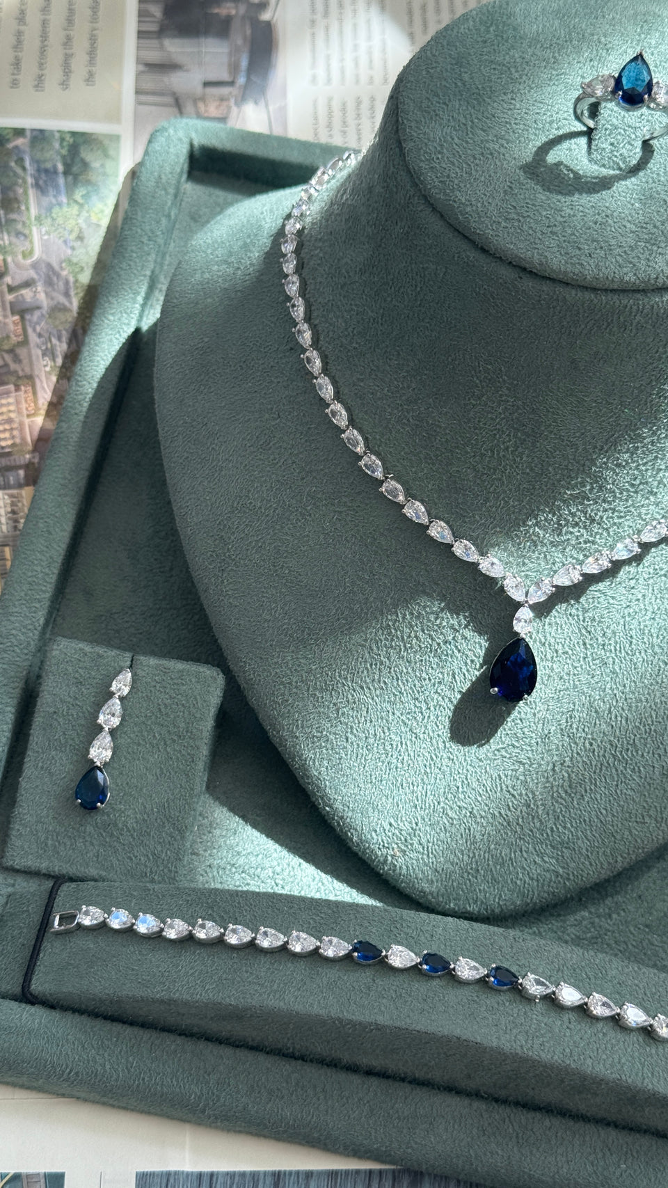 A jewelry set displayed on a green velvet jewelry bust, featuring a silver-tone necklace with a teardrop-shaped dark blue gemstone, a matching bracelet, a matching ring, and a small matching earring.