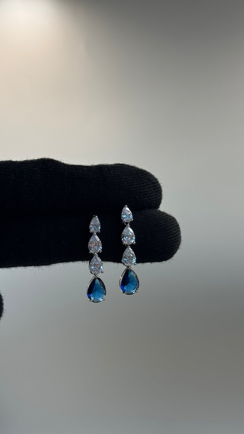 A pair of silver-tone drop earrings with clear marquise-cut stones and blue teardrop-shaped stones, held by a black gloved hand against a neutral gray background.