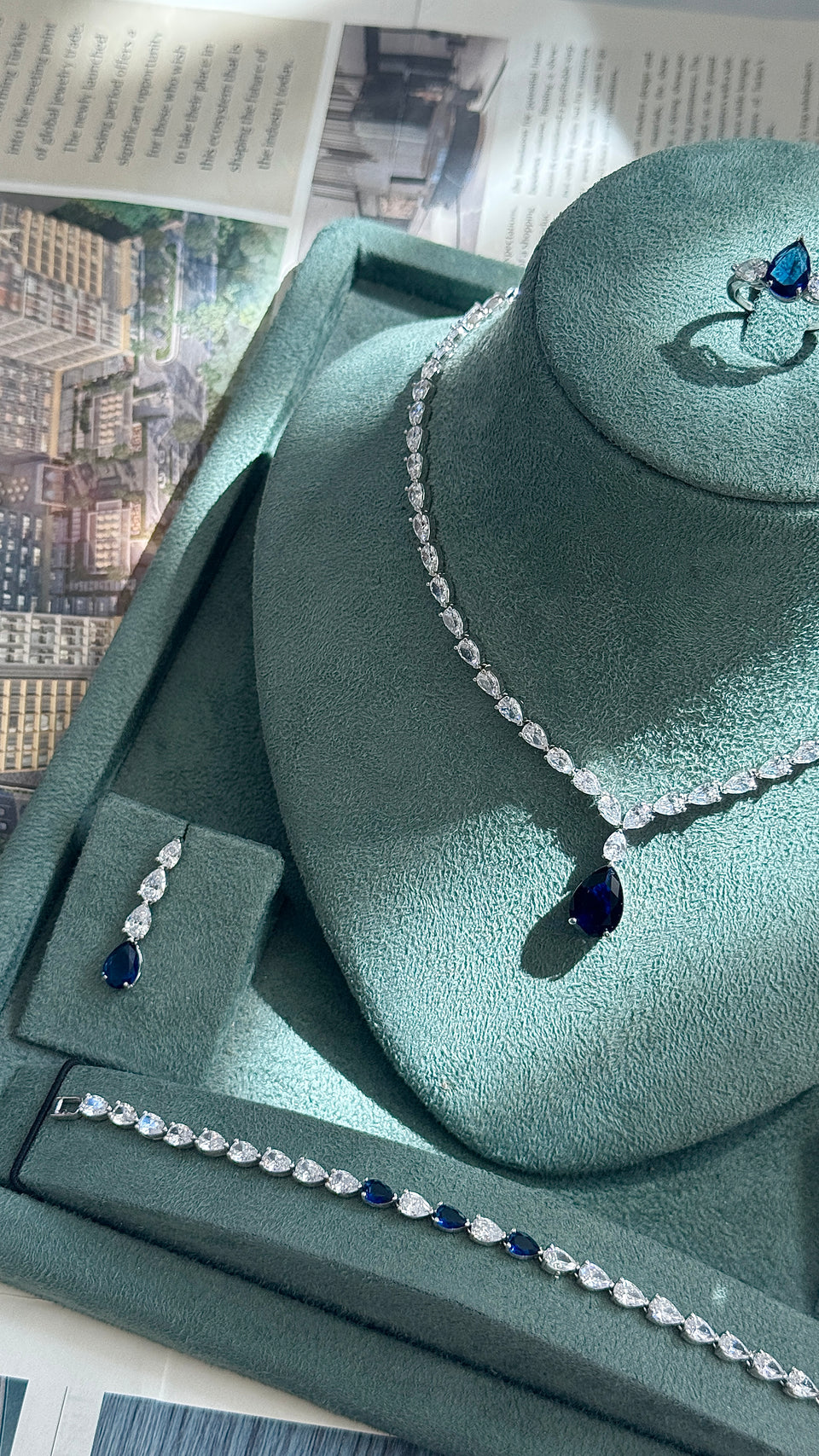 A jewelry set displayed on a green velvet jewelry bust, including a necklace with a teardrop-shaped dark blue stone, matching drop earrings, and a bracelet, arranged on a green presentation tray with printed papers in the background.