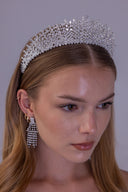 A woman wearing a sparkling silver rhinestone tiara and matching dangling rhinestone earrings against a plain gray studio background.