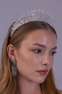 A woman wearing a sparkling silver rhinestone tiara and matching dangling rhinestone earrings against a plain gray background.
