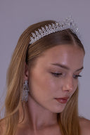 A woman wearing a sparkling silver tiara headband and matching dangling rhinestone earrings, shown in a close-up portrait against a plain light gray background.