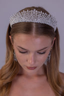 A woman wearing a sparkling silver rhinestone tiara and matching drop earrings, looking down against a plain light gray background.
